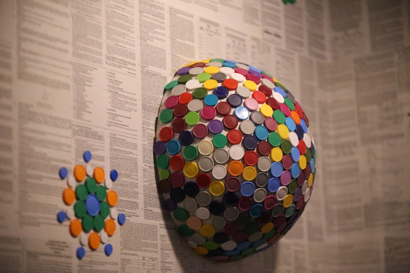 A dome covered in colorful bottle caps mounted on a wall of newspaper